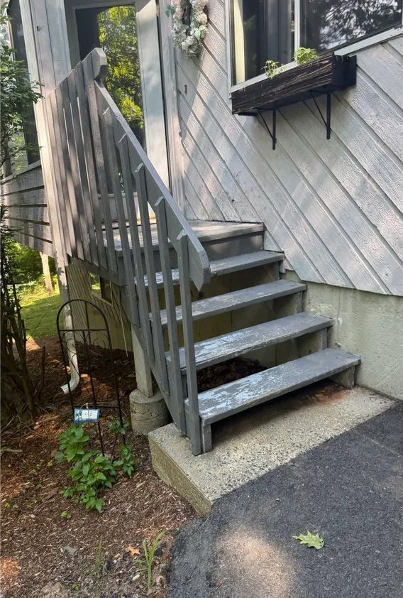 Gray wooden staircase leading up to a light gray building with a window box. Concrete steps at the base.