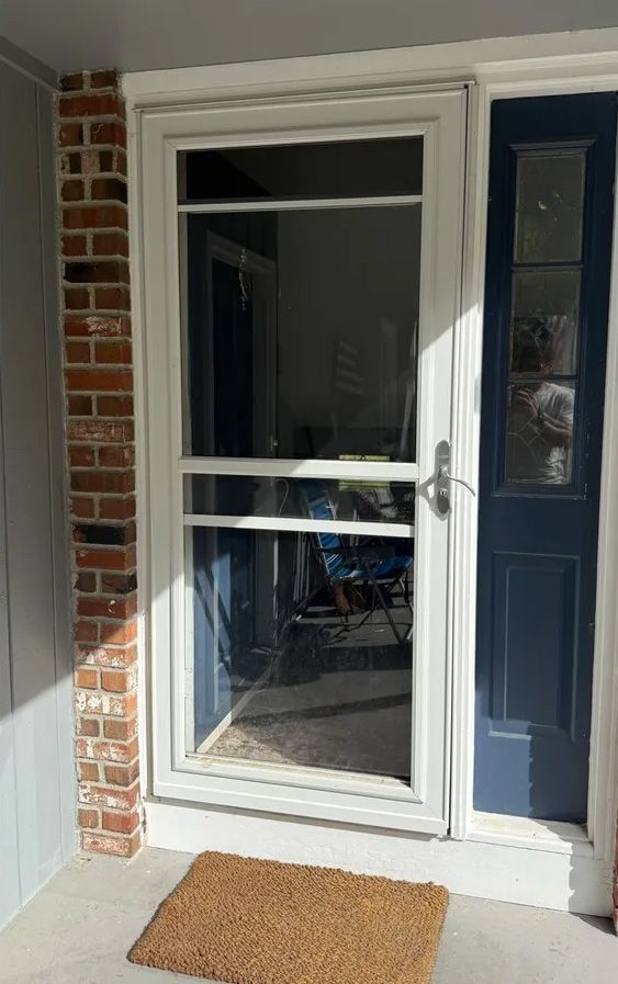 White screen door with a glass panel, brick facade, and navy door. Brown welcome mat.