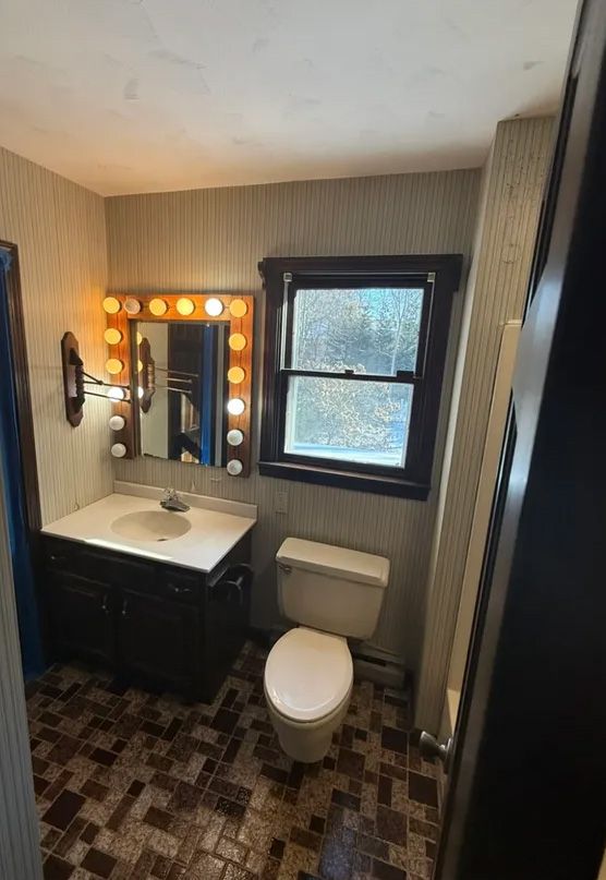 Bathroom with a dark vanity, lit mirror, toilet, window, and patterned floor.