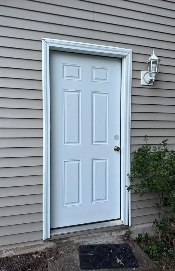 White exterior door with six panels, a light fixture, and a welcome mat on gray siding.