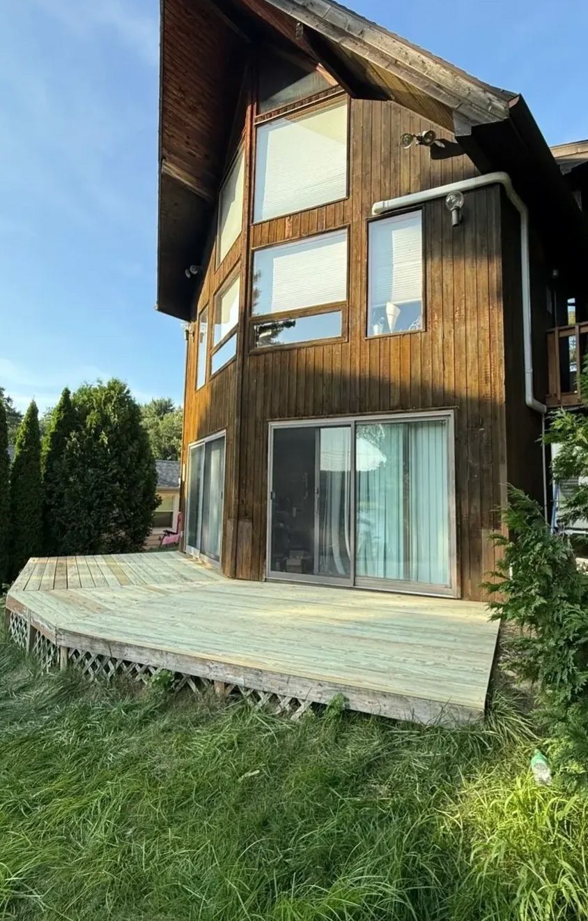 Wooden house with a deck and large windows on a grassy hill, under a blue sky.