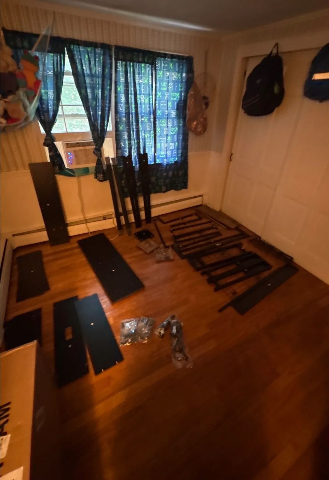 Disassembled furniture pieces scattered on a hardwood floor in a room with a window, curtains, and a closed door.