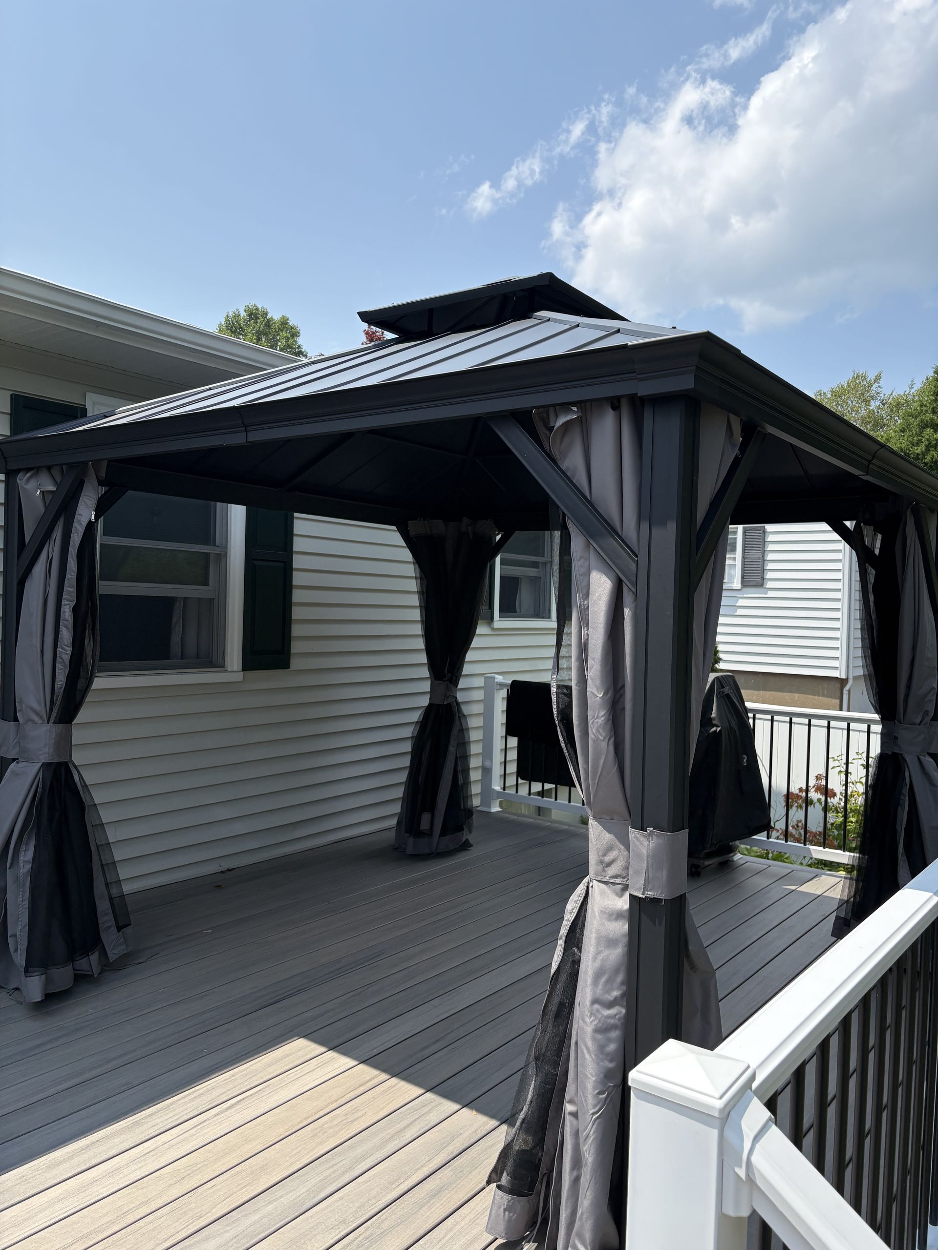 Black gazebo with gray curtains on a wooden deck, next to a white house with black shutters, under a partly cloudy sky.