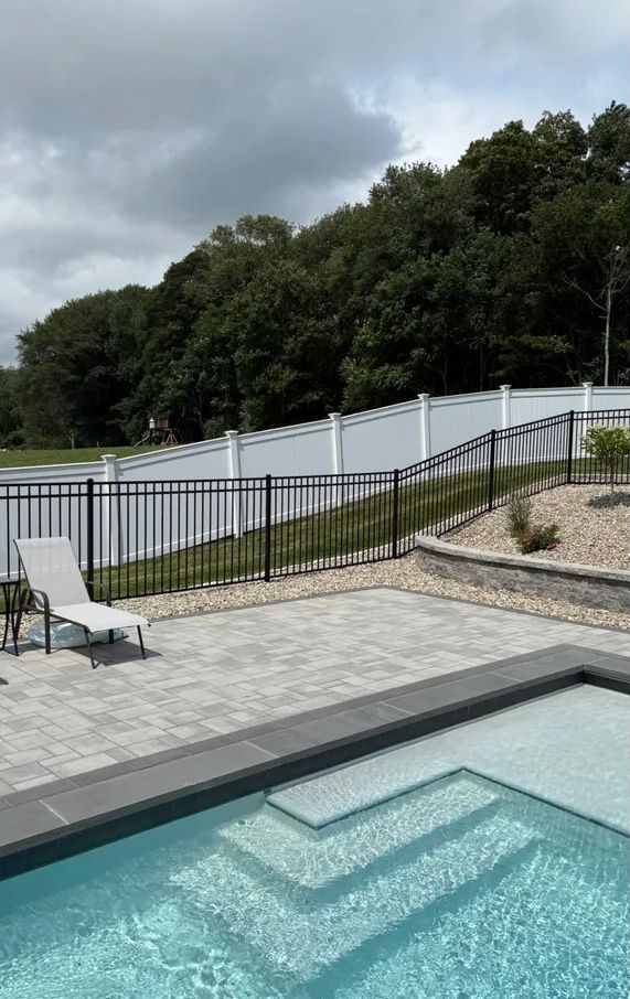 Poolside scene with a pool, patio, white and black fence, and trees.