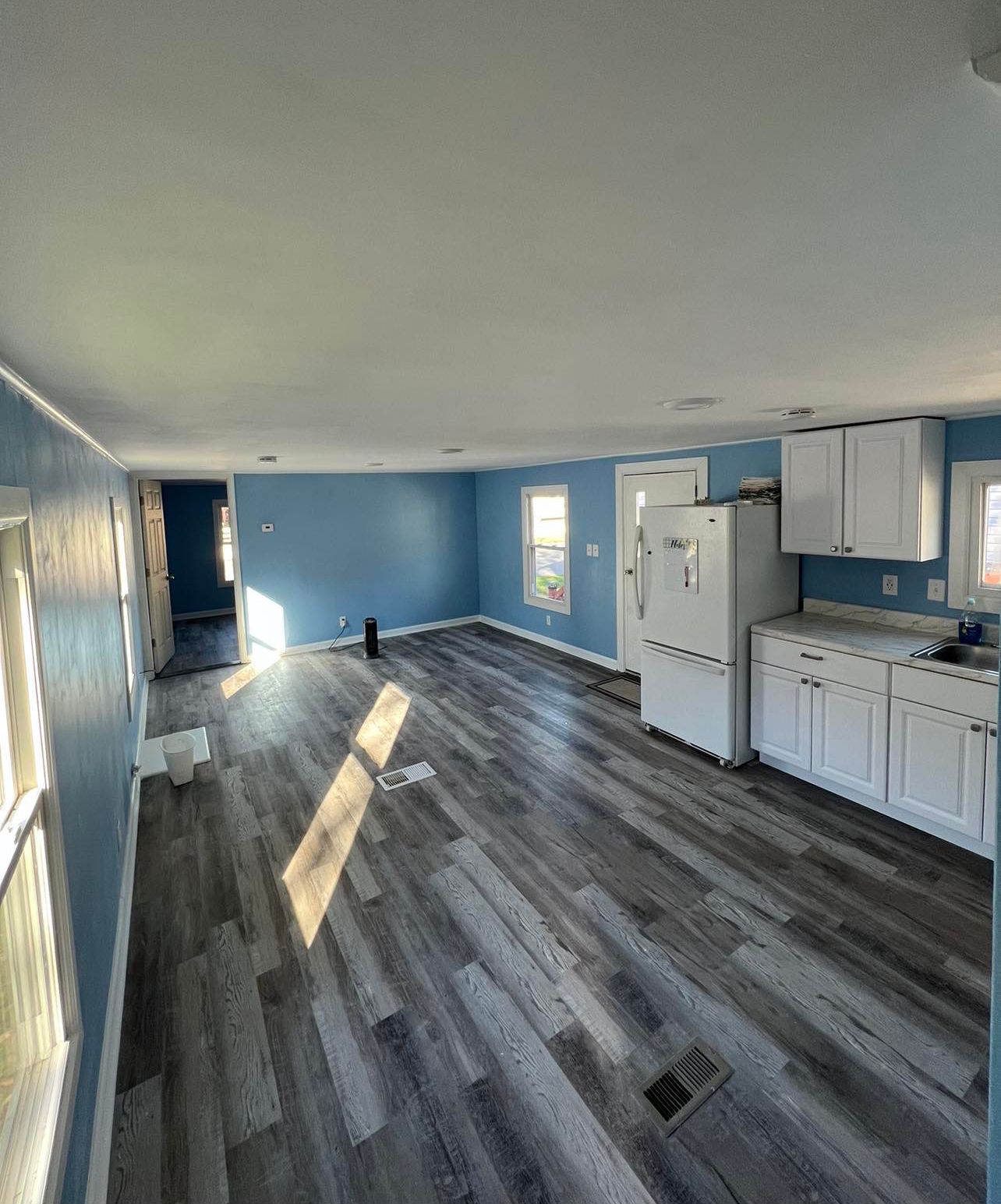 Blue-walled interior with gray flooring, white cabinets, refrigerator, and doorway. Sunlight streams through the windows.