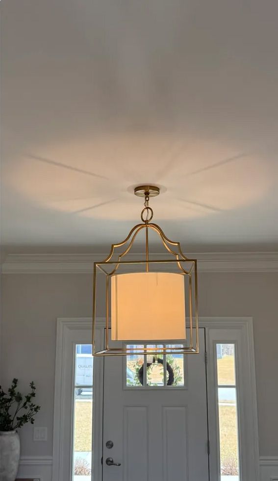 Gold framed pendant light fixture hanging from a white ceiling, casting a shadow above a front door with a wreath.