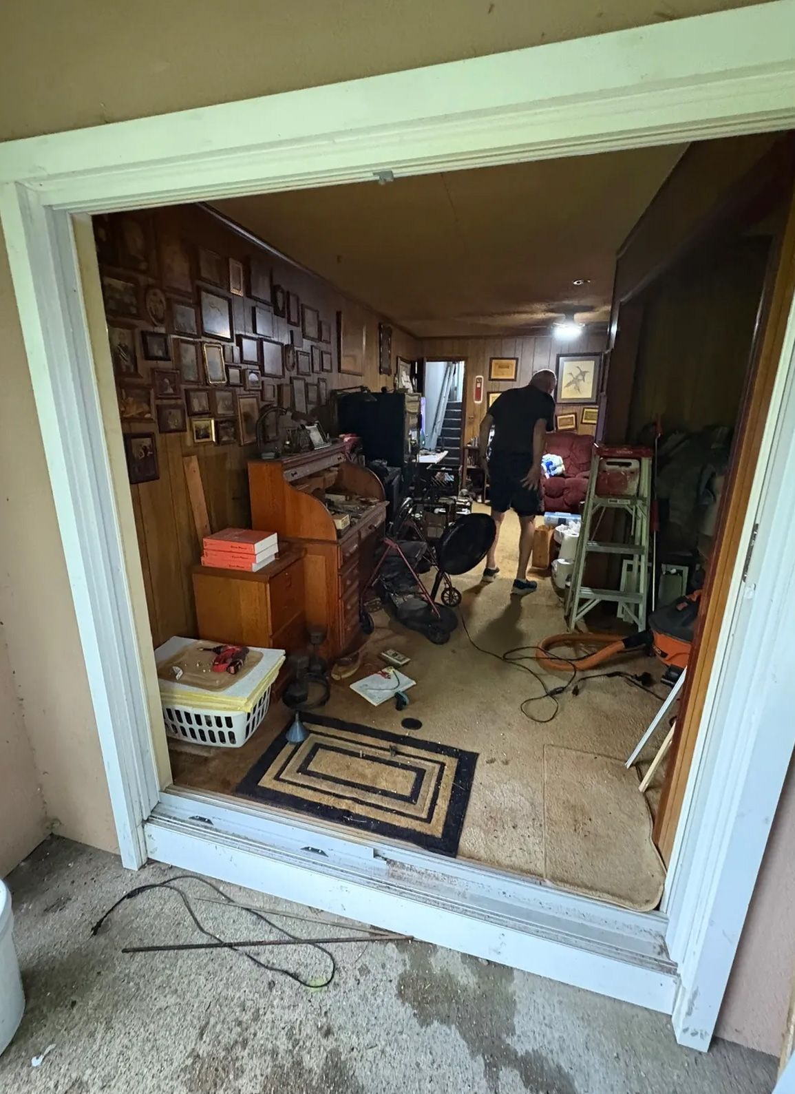View from doorway into cluttered room; person inside, dark walls, various items and furniture.