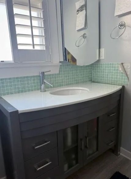 Gray vanity with white countertop and sink, light green tile backsplash, and cabinet above.