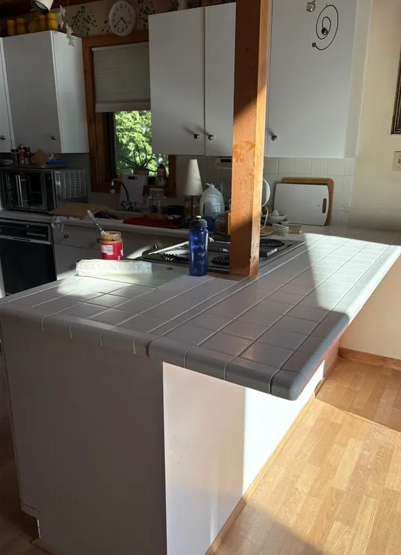 Kitchen with white cabinets, light gray tile countertop, wooden post, and a sunny window.
