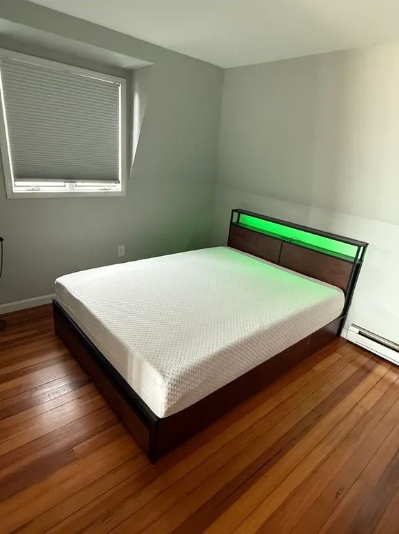 Bed with a green-lit headboard, white mattress, in a room with a window, wood floor, and gray walls.