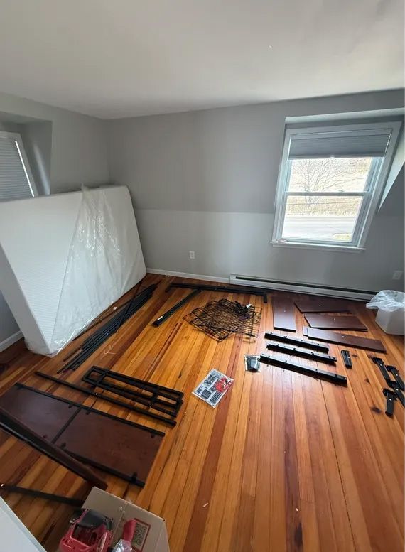 Bedroom with unassembled bed parts on a hardwood floor, mattress leaning against the wall.