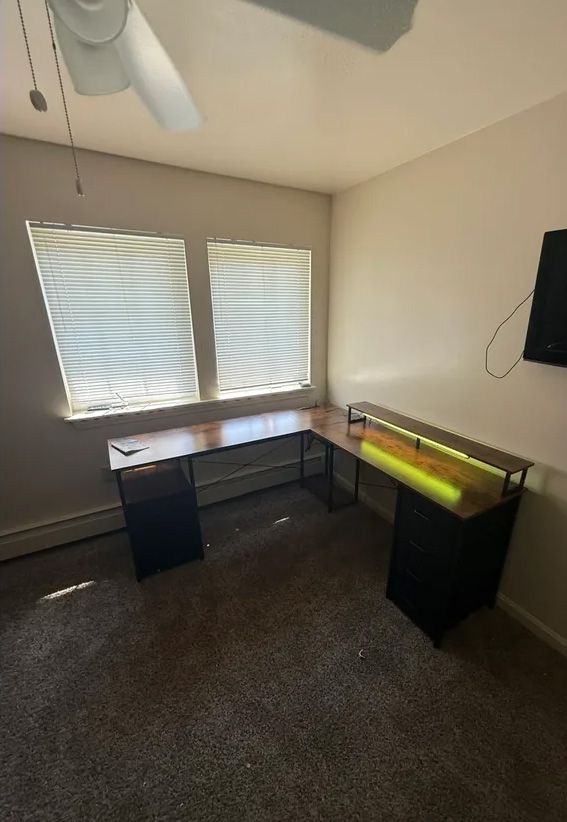 L-shaped desk in a room with a window, carpet, and a ceiling fan; the desk has a black frame, wood top, and a yellow light.