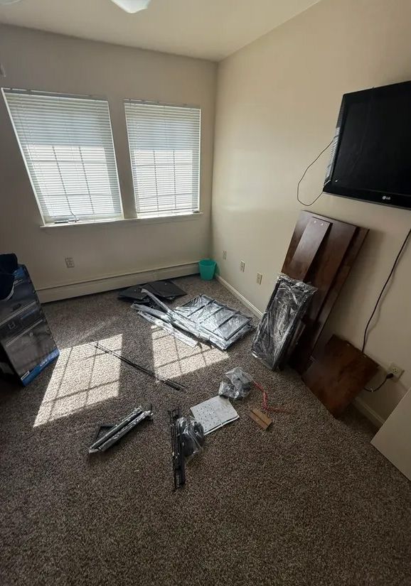 A room with disassembled furniture on the floor. Windows, a TV on the wall, and carpet are visible.