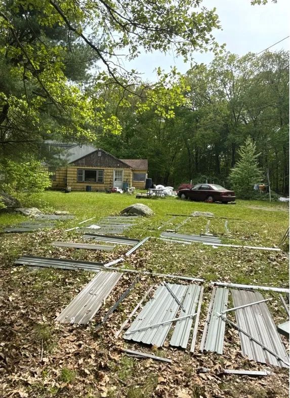 A house with a car in a yard, debris scattered across the grass, surrounded by trees.
