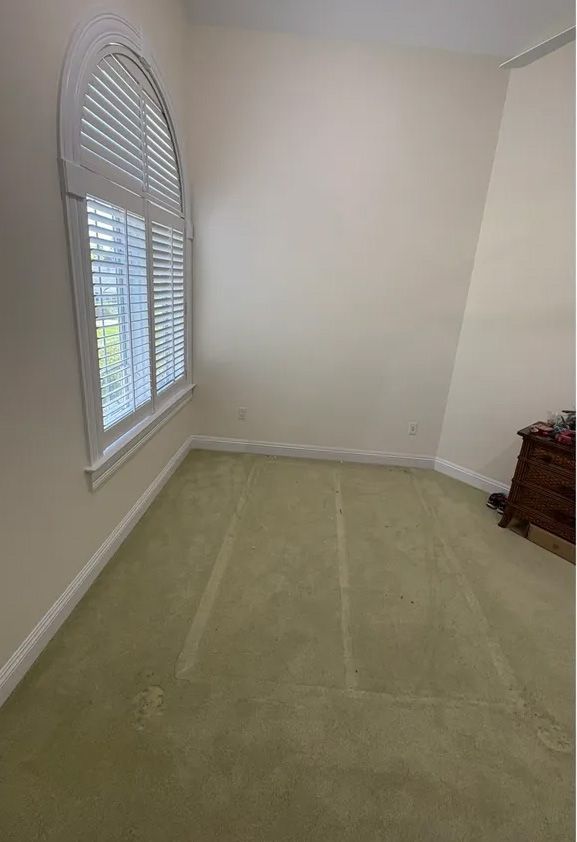 Room with arched window and green carpet. Beige walls, small wooden table in corner.
