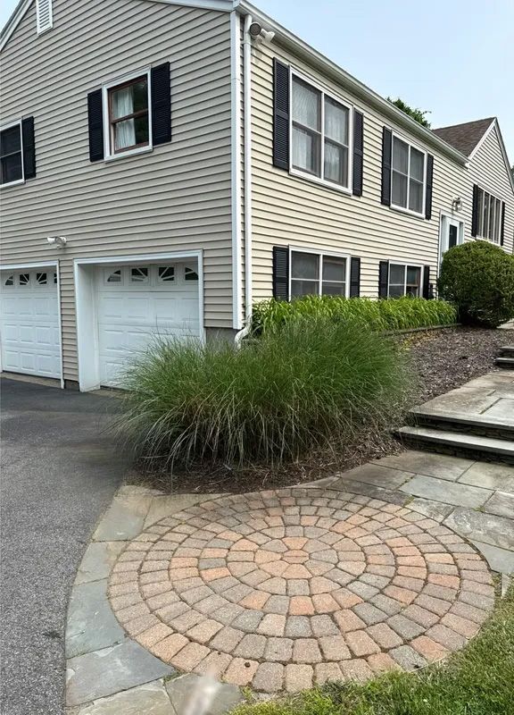 Two-story house with tan siding, black shutters, and a circular brick patio. Tall green ornamental grass in foreground.