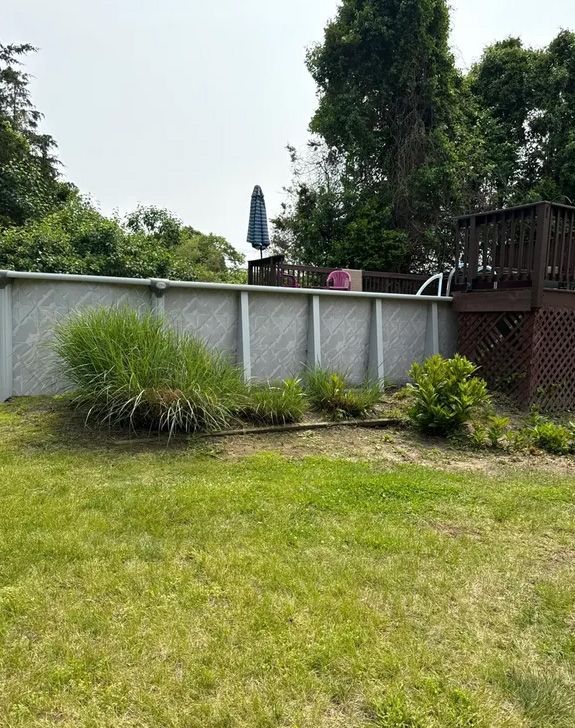 Backyard view with above-ground pool, plants, lawn, and wooden deck under cloudy sky.