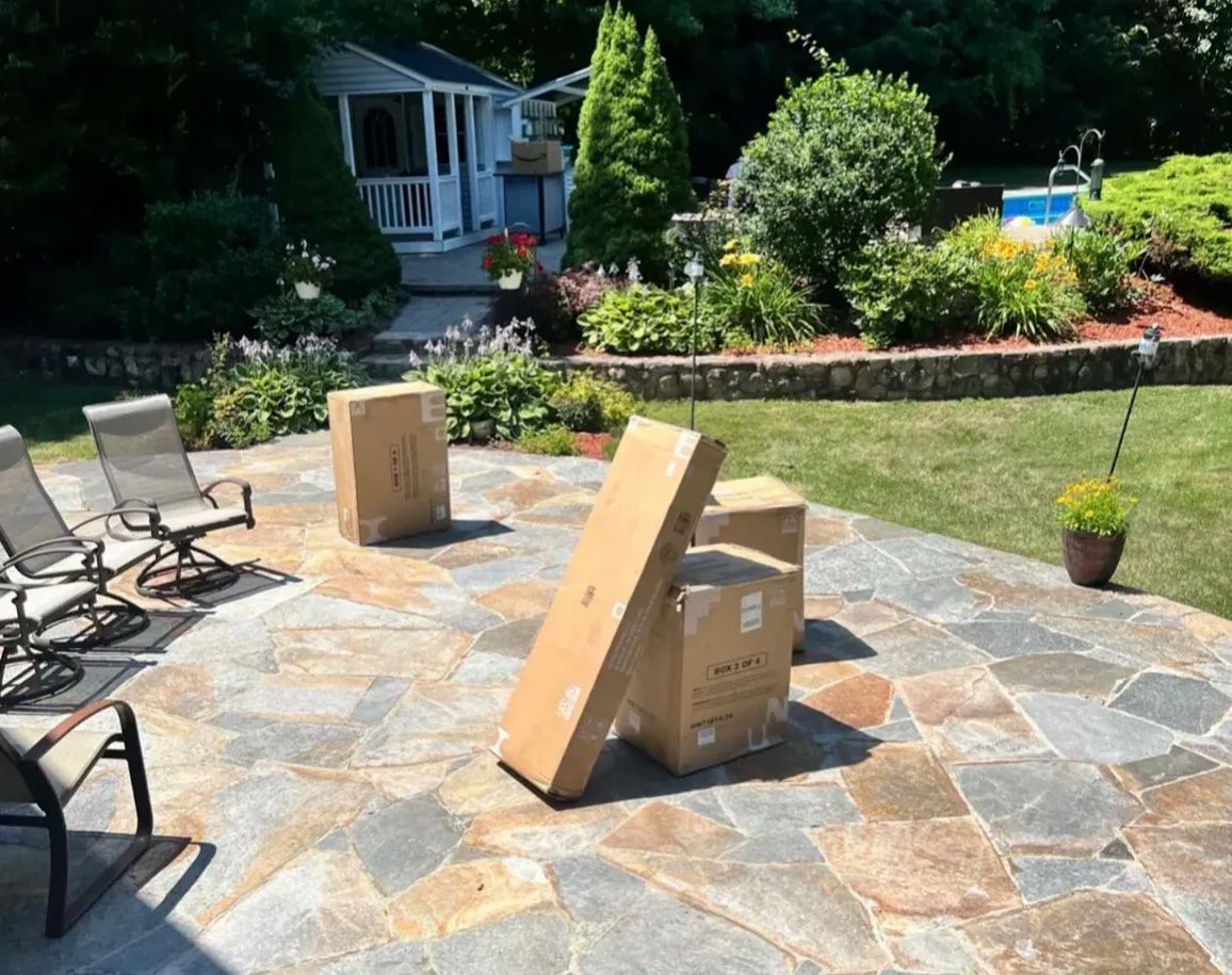 Cardboard boxes on a stone patio in a backyard with chairs, greenery, and a shed.
