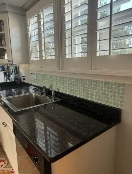 Kitchen counter with black granite, stainless steel sink, and light green tile backsplash. White cabinets and window shutters.