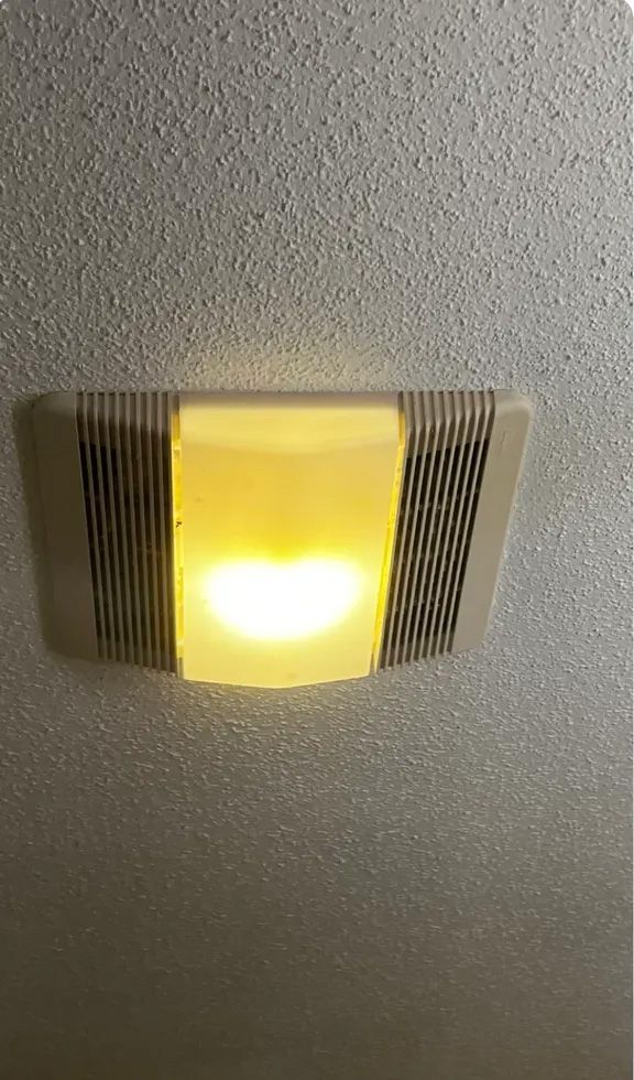 Bright yellow light shines from a rectangular wall-mounted fixture with vertical black stripes on a textured, light gray surface.