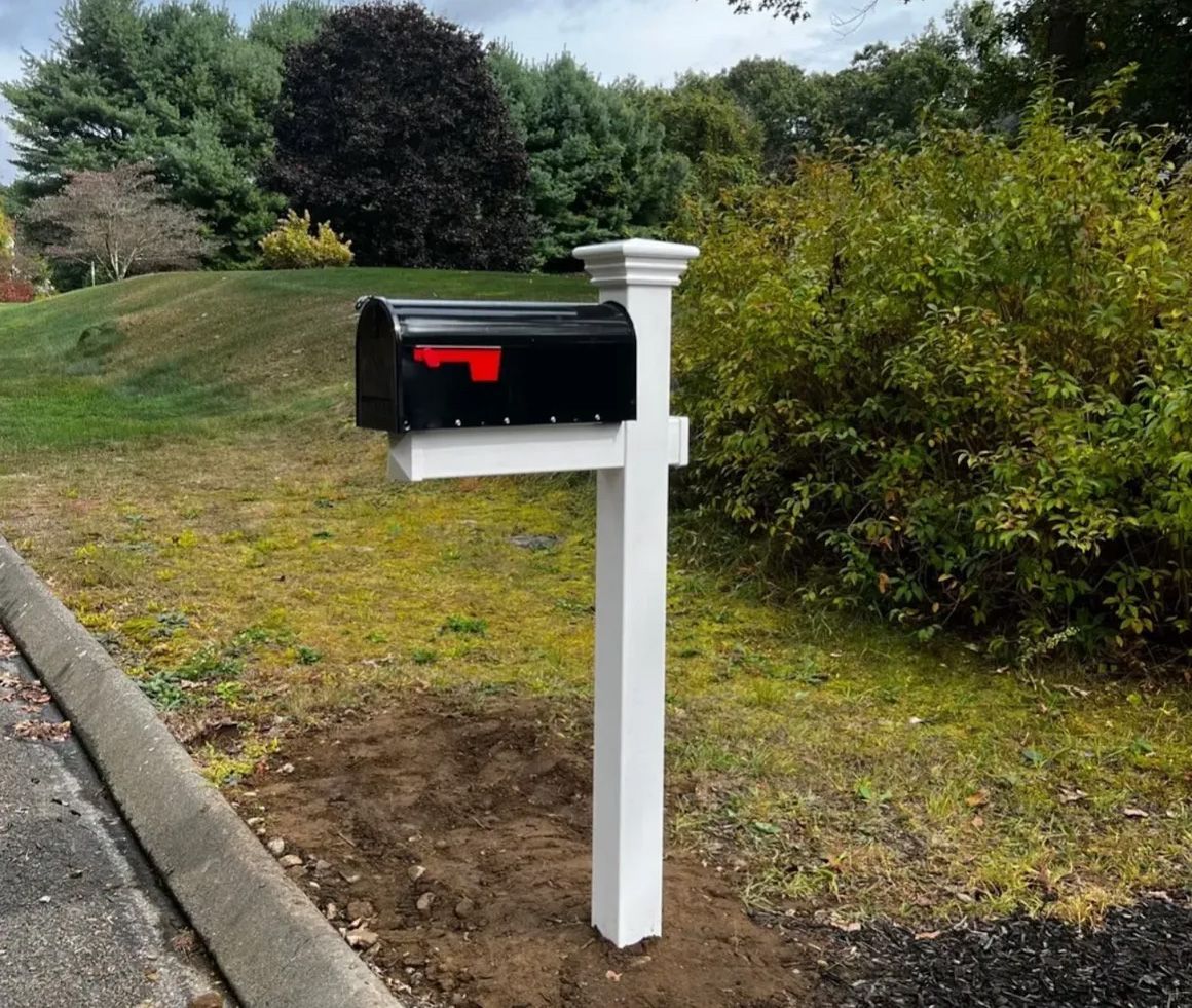 Black mailbox with red flag on white post next to a road, set against trees and a grassy hill.