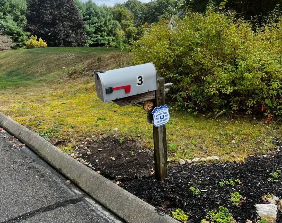 Gray mailbox with number 3 and red flag, next to asphalt road and landscaped yard.