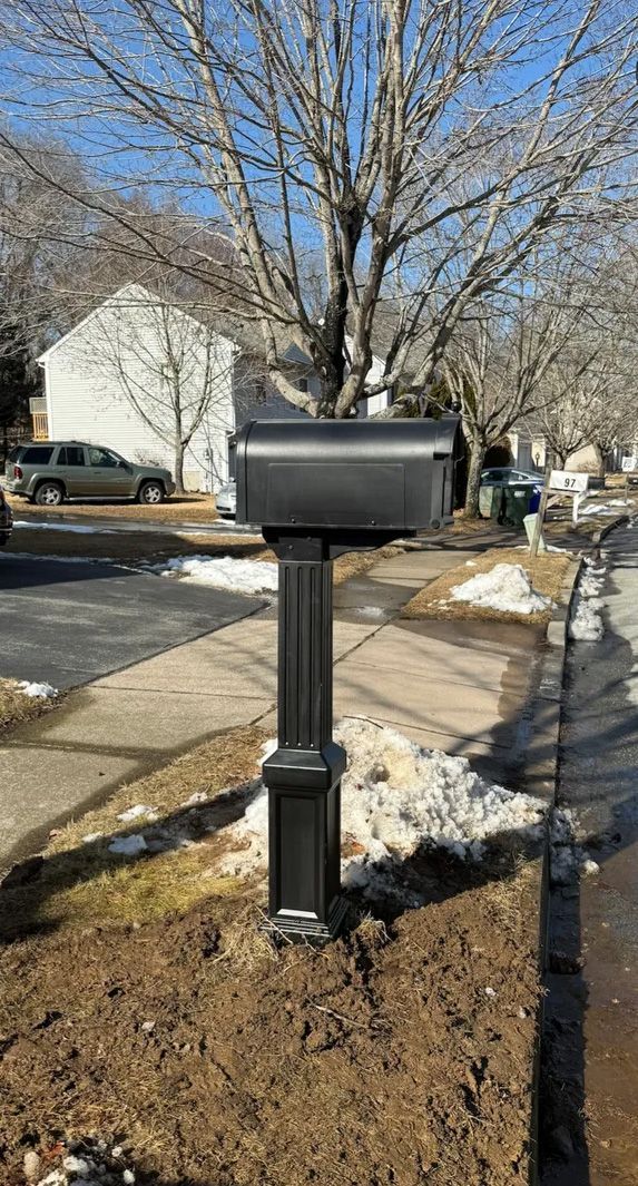 Black mailbox on a black post next to a sidewalk with snow. Background includes houses and a tree.