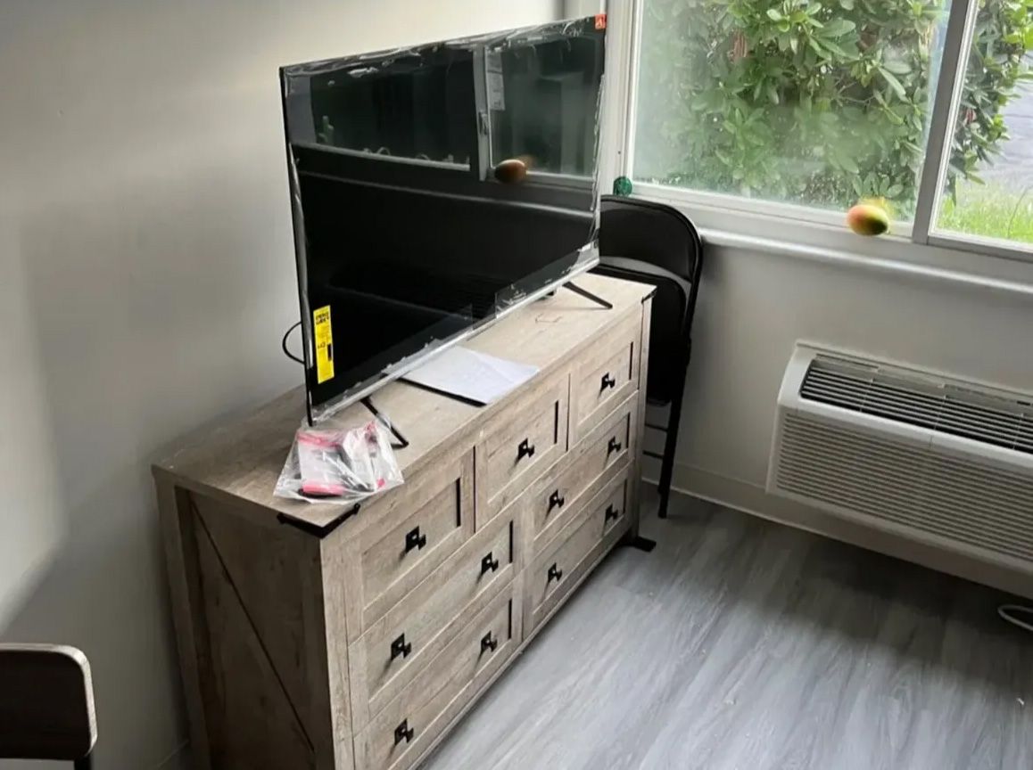 A TV sits atop a wooden dresser in front of a window. A chair and an air vent are also visible.