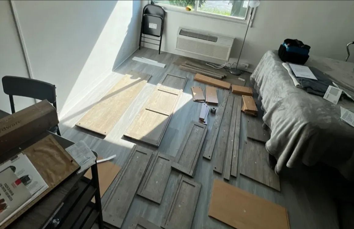 Disassembled furniture pieces scattered on a floor, near a bed and a window in a room.