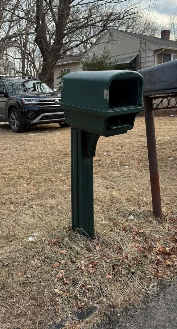 Green mailbox on a post in front of a house, car parked in the background, daytime.