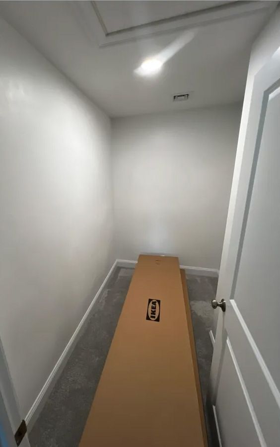 Small, empty room with a cardboard box on the floor; white walls, door, and a ceiling access point.