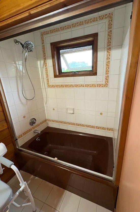 Brown-toned bathtub/shower stall with white tiles, glass door, window, and a shower head.