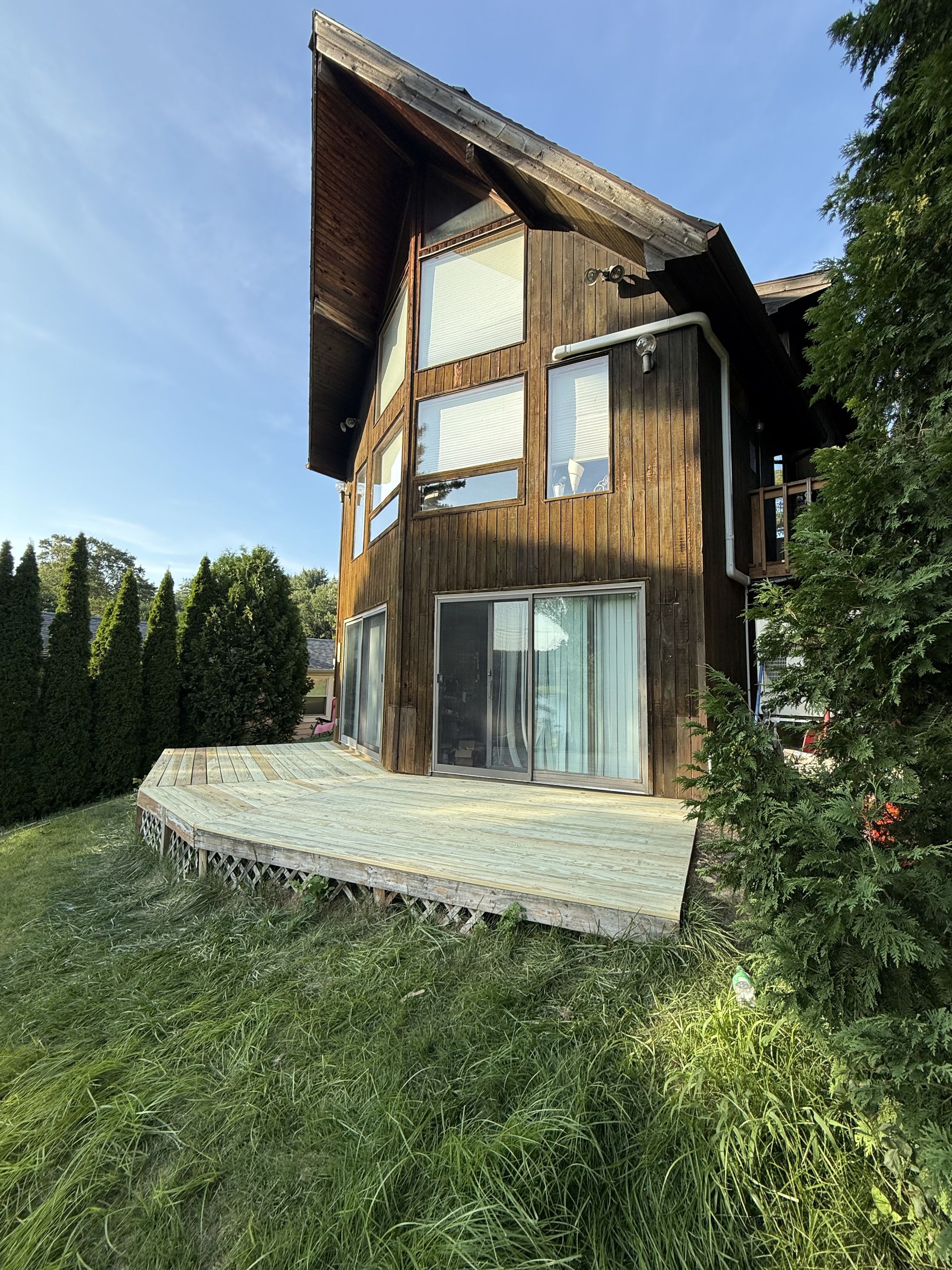 Wooden house with a sloping roof, windows, and a patio, surrounded by trees and grass.