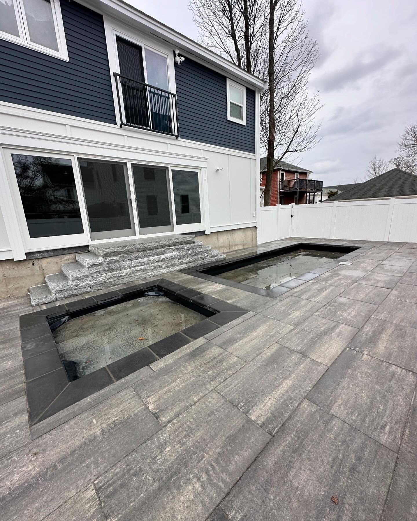 Backyard patio with two rectangular in-ground pools and a two-story blue and white house. Gray pavers and steps.