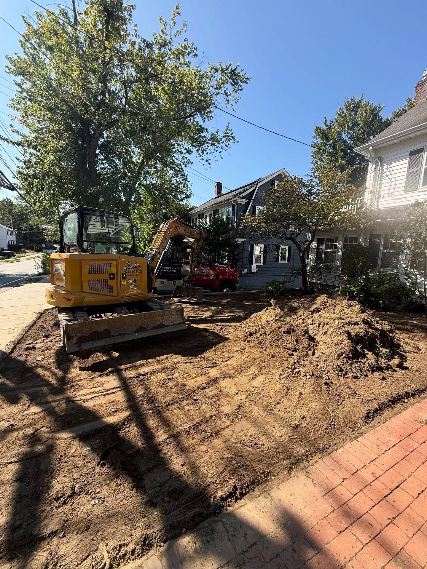 Yellow excavator on dirt, clearing a yard next to a blue house and red brick sidewalk under a blue sky.