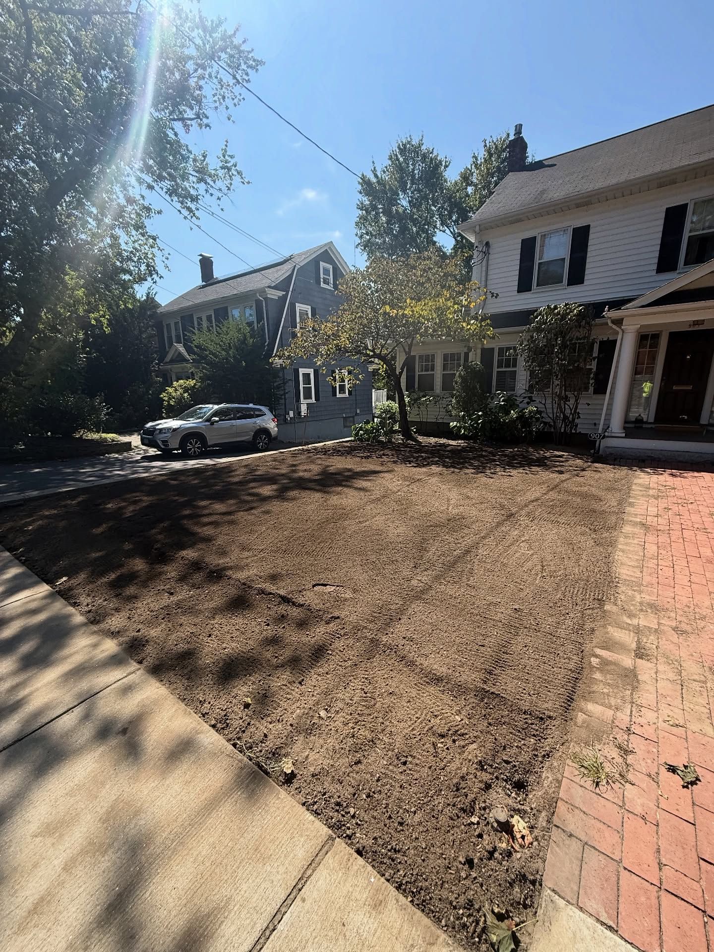 Dirt patch in front yard, brick sidewalk, trees, houses, car, sunny day.