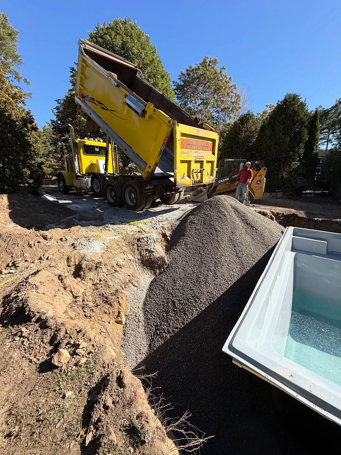 Yellow dump truck unloading gravel into a trench next to a pool shell. A person stands nearby.
