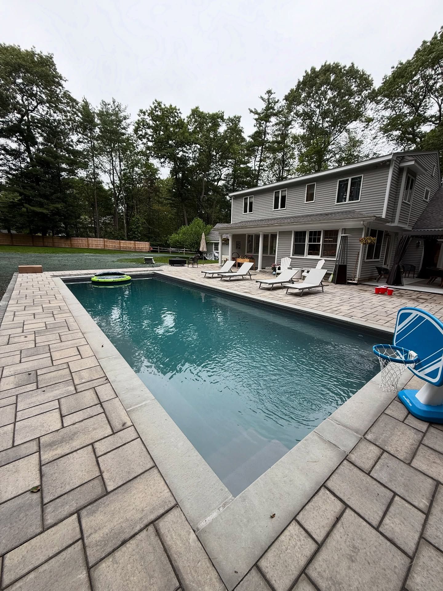 Rectangular pool with gray pavers, white lounge chairs, and a two-story house in the background.