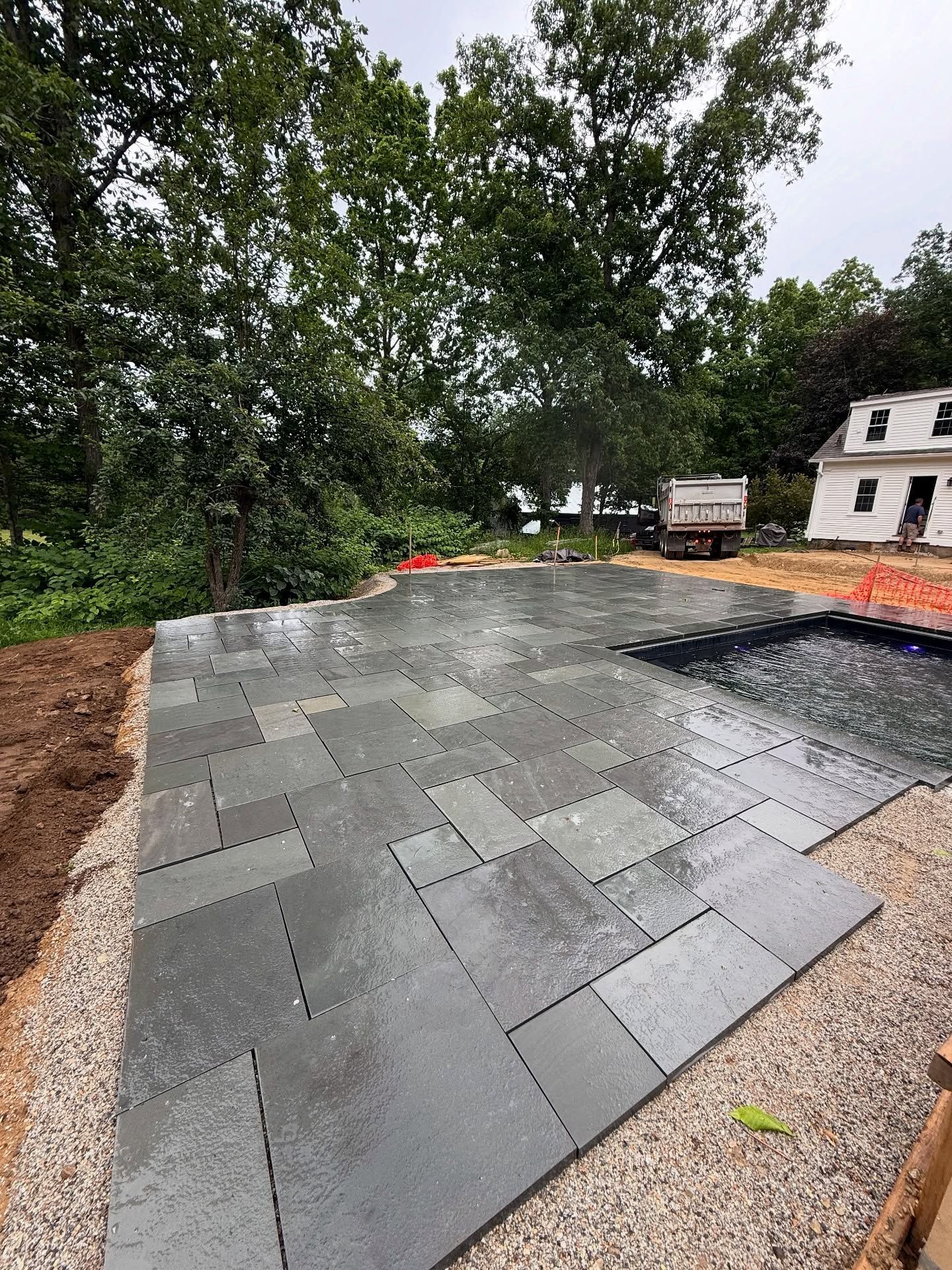 New blue stone patio being installed, surrounded by gravel and soil. Green trees and a white house in the background.