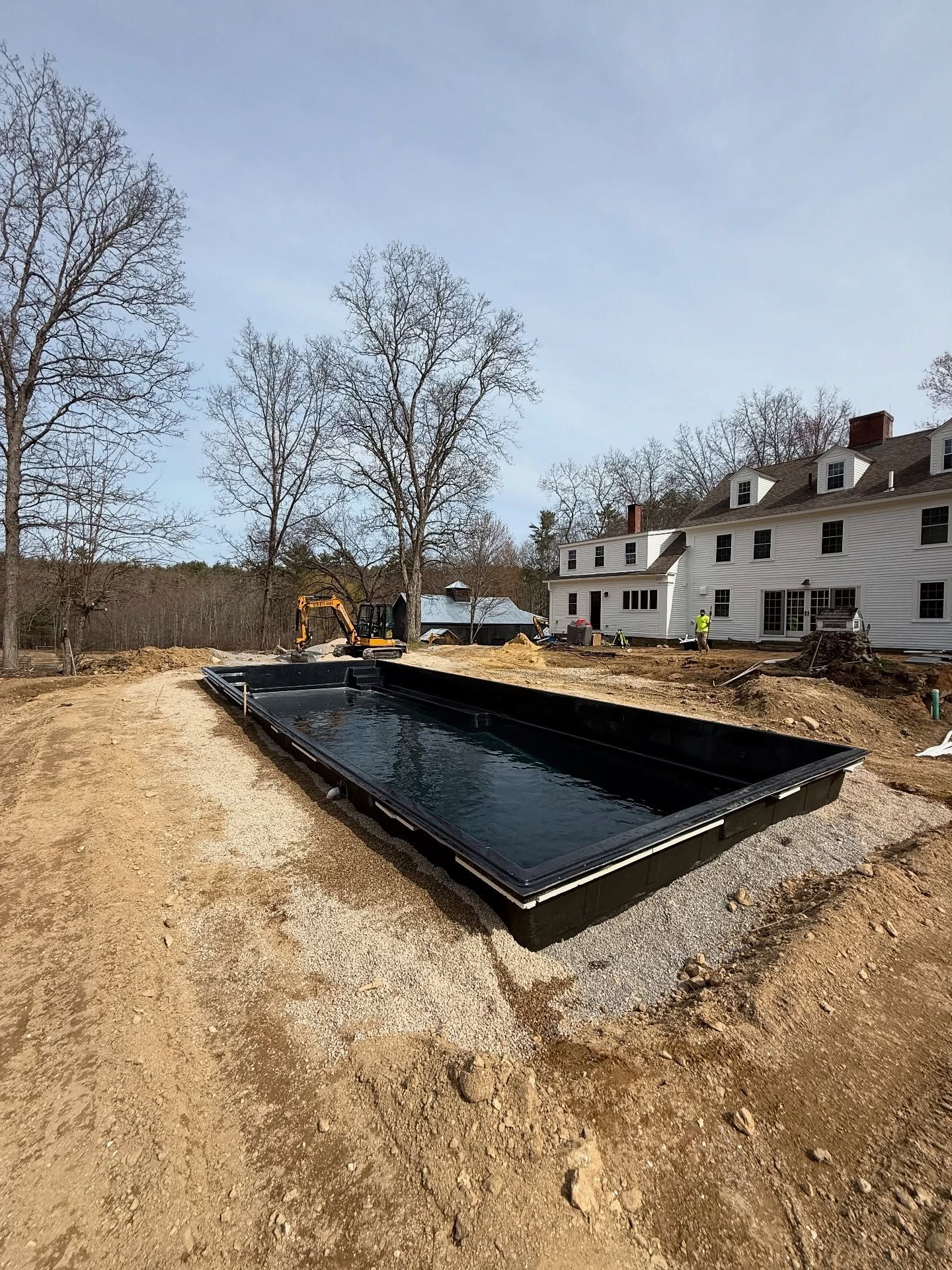 Rectangular black swimming pool shell in a dirt lot, near a white building with red chimneys.