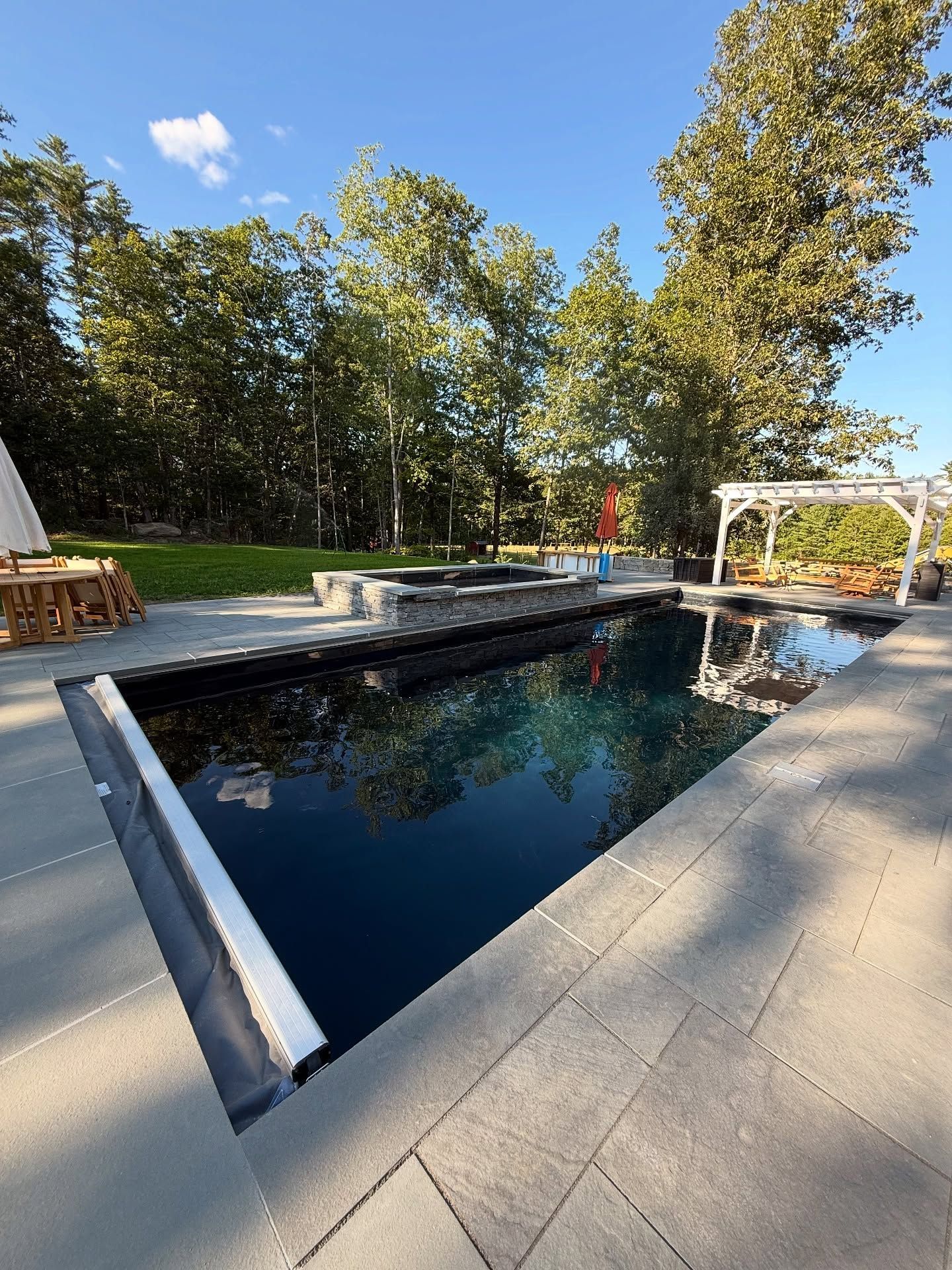 Dark-bottomed rectangular pool reflects sky, adjacent to a patio with a jacuzzi, gazebo, and surrounding trees.