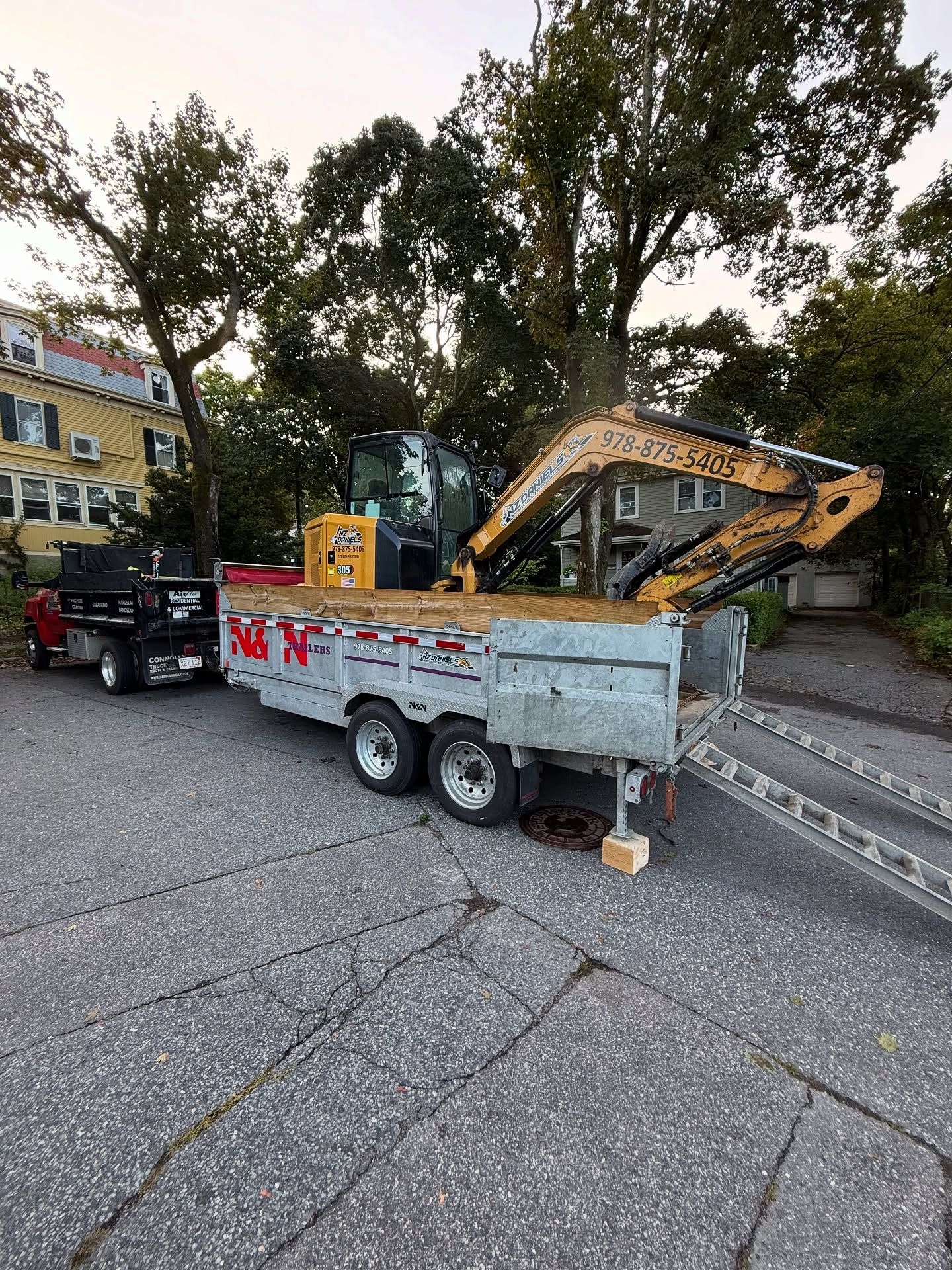 Excavator on a trailer being towed by a pickup truck, parked on pavement, with buildings and trees in background.