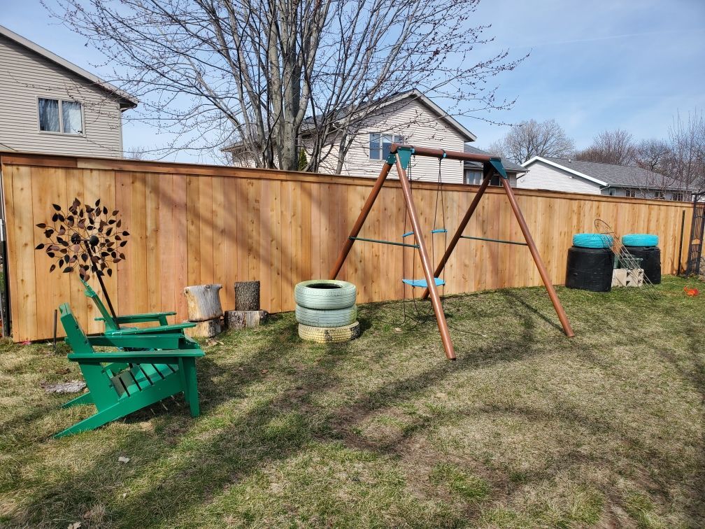 A wooden fence with a swing set and a green chair in the backyard.