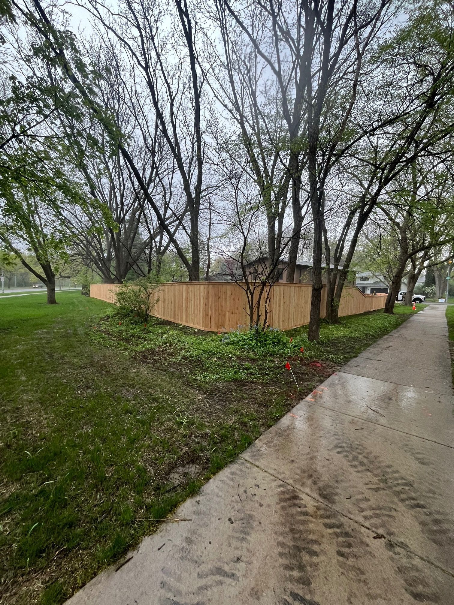 A wooden fence is surrounded by trees on the side of a sidewalk.