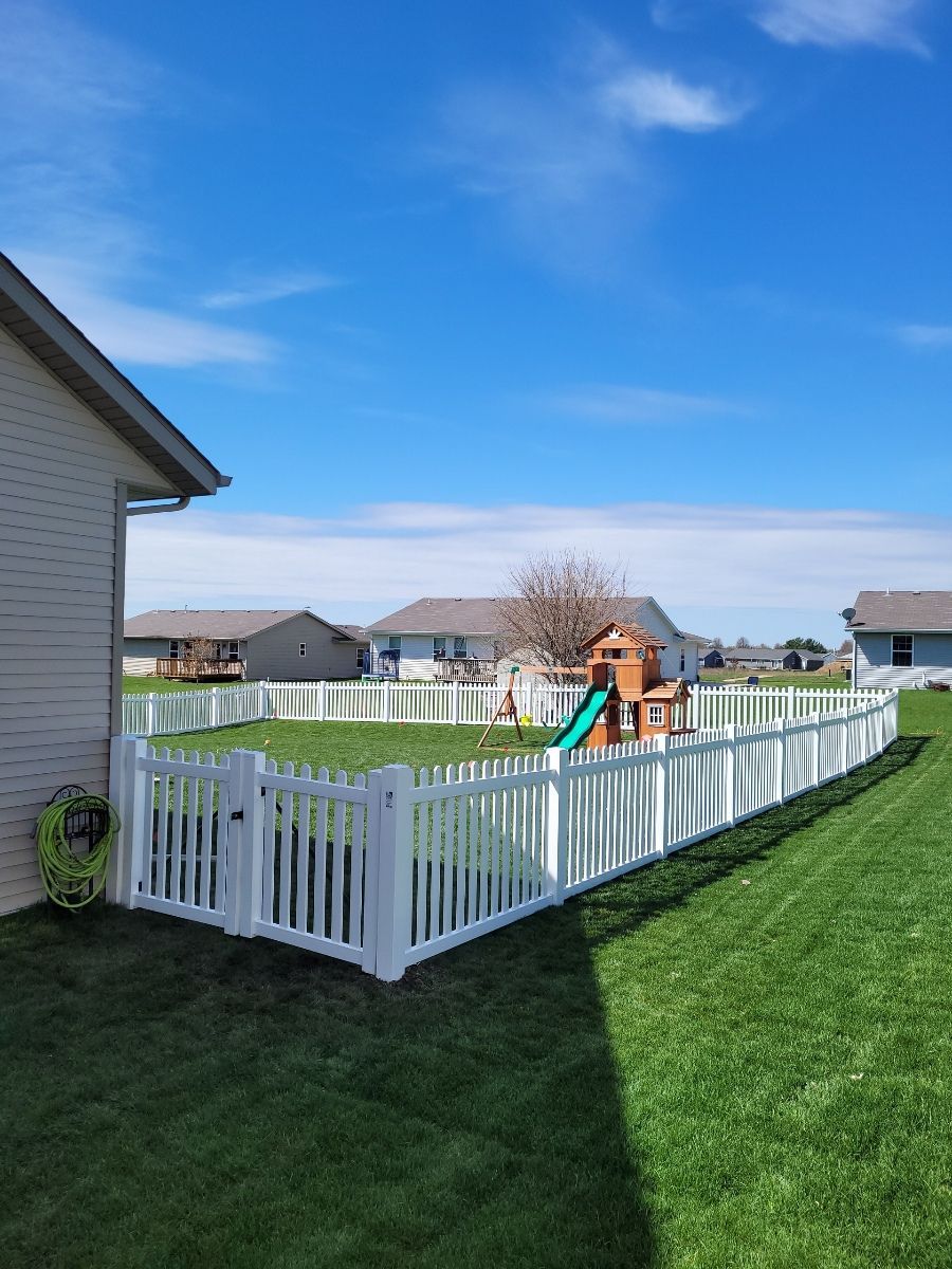 A white picket fence surrounds a backyard with a playground in the background.