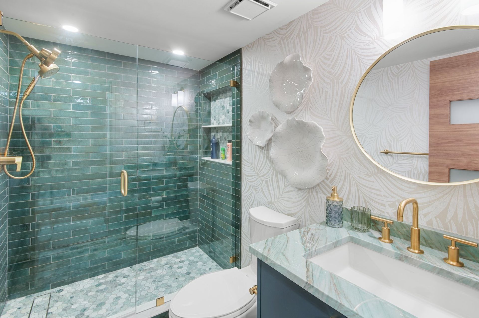 Bathroom with teal tiled shower, gold fixtures, and textured wallpaper with white floral accents.