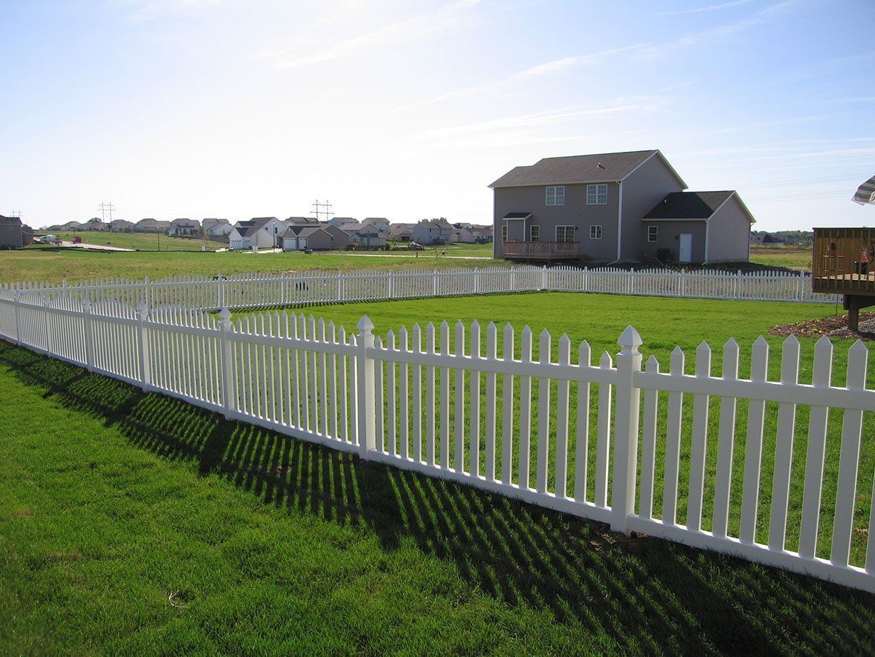 A white picket fence surrounds a lush green field with a house in the background