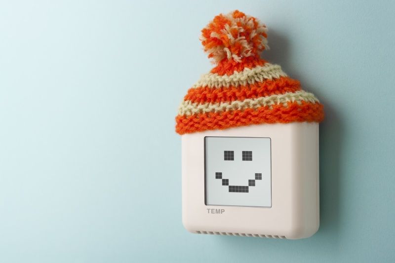 Thermostat with a smiley face display, wearing an orange and cream knitted hat, mounted on a light blue wall.