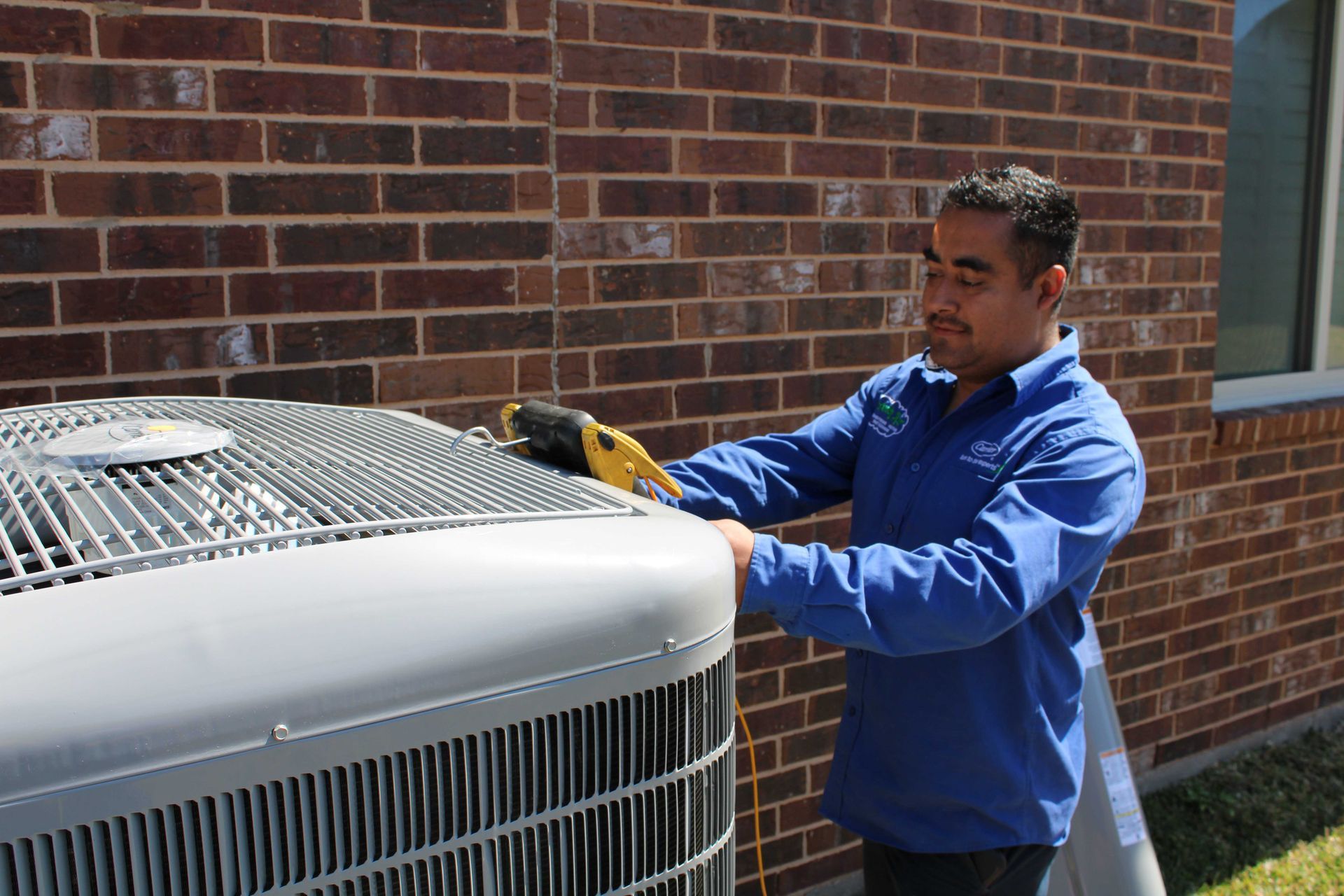 HVAC technician working on an air conditioning unit near a brick wall, using a tool.
