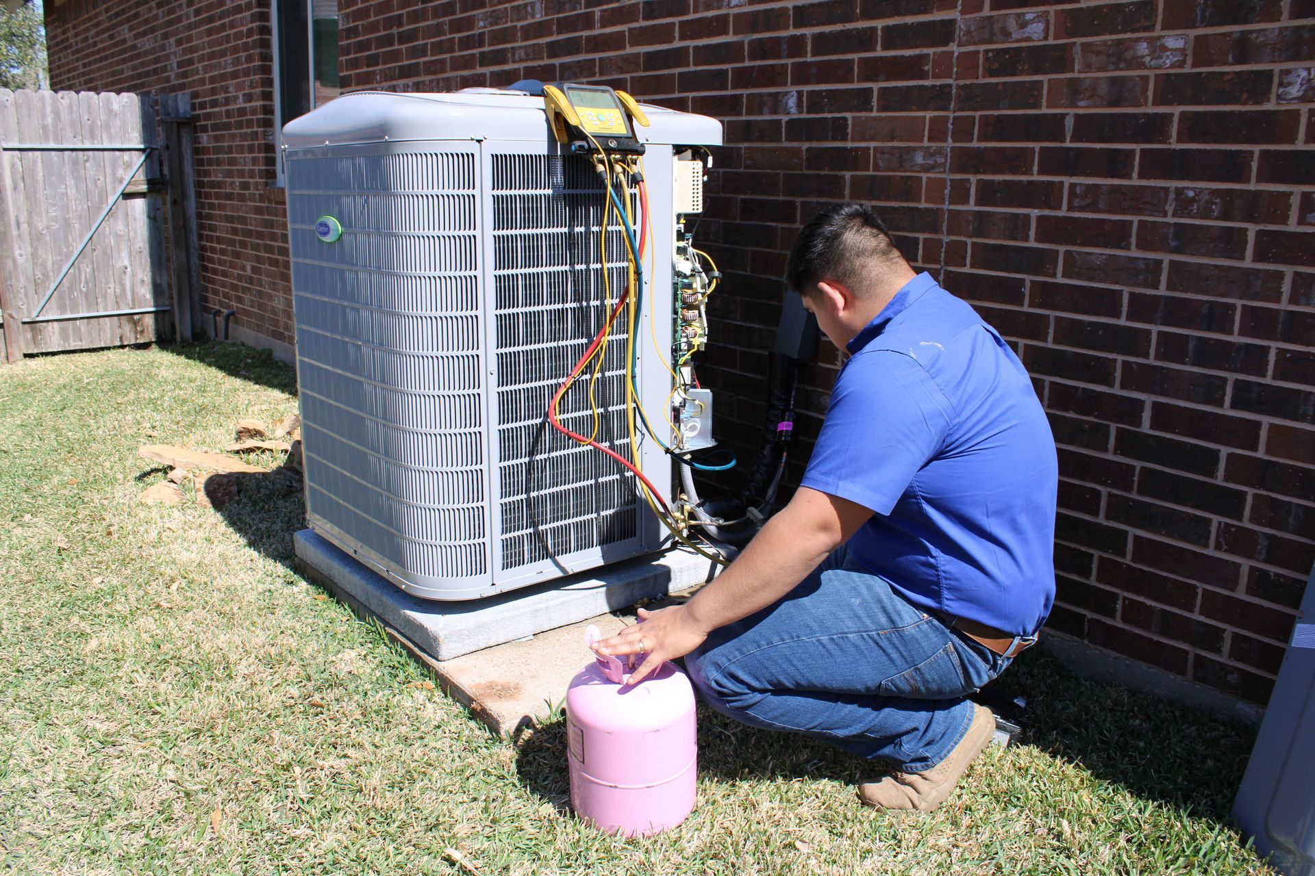 HVAC technician repairs an outdoor AC unit; he kneels with a pink refrigerant tank next to the unit.